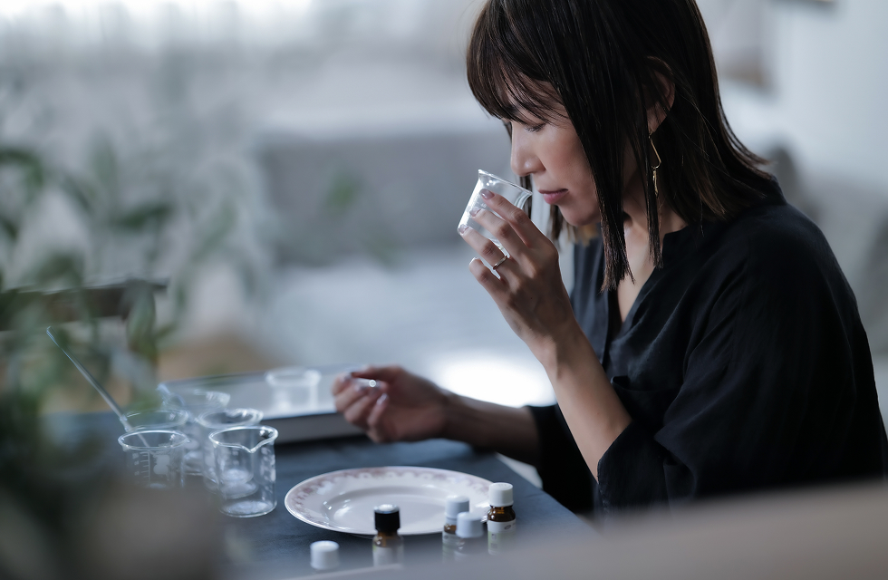 女性がビーカーから香りを嗅いでいる様子 机の上にはたくさんのガラスビーカーと精油