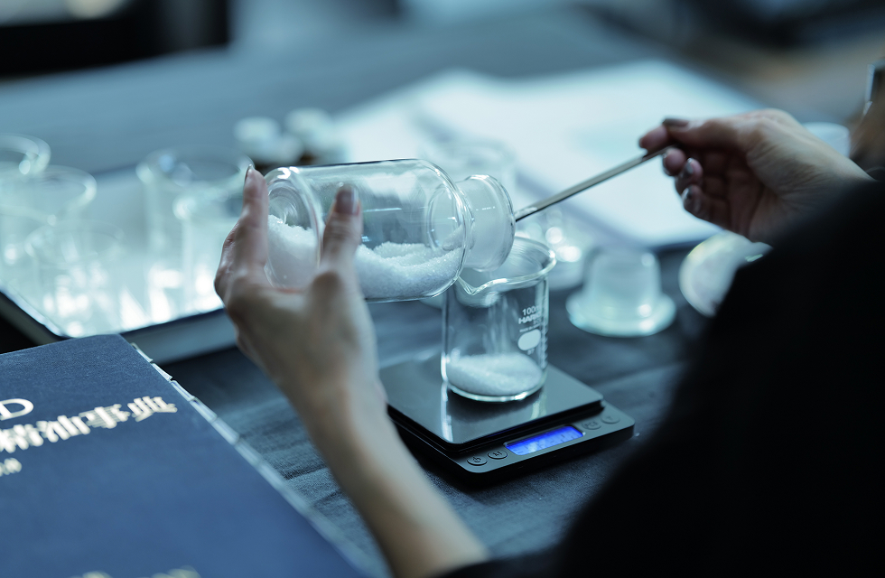 ガラスのビーカーに粉を測っているところ。机の上にはたくさんのガラスのビーカーと精油辞典