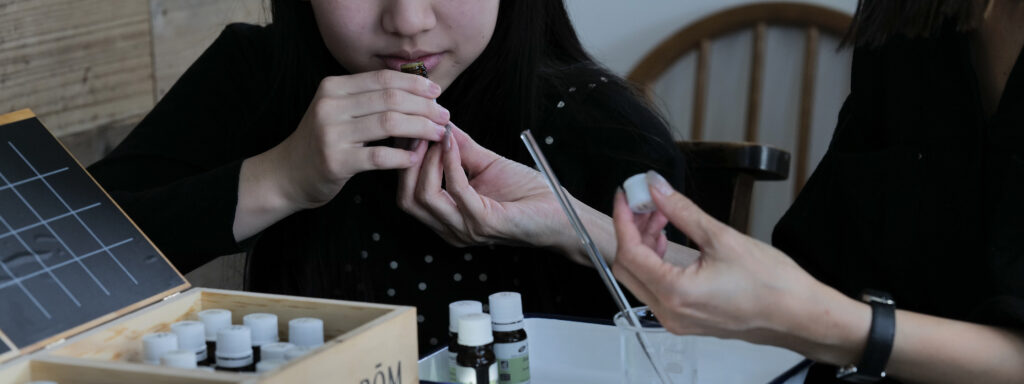 女の子に精油を差し出している。女の子はその精油の香りを嗅いでいる。テーブルの上にはたくさんの精油やガラスのビーカーが置いてある