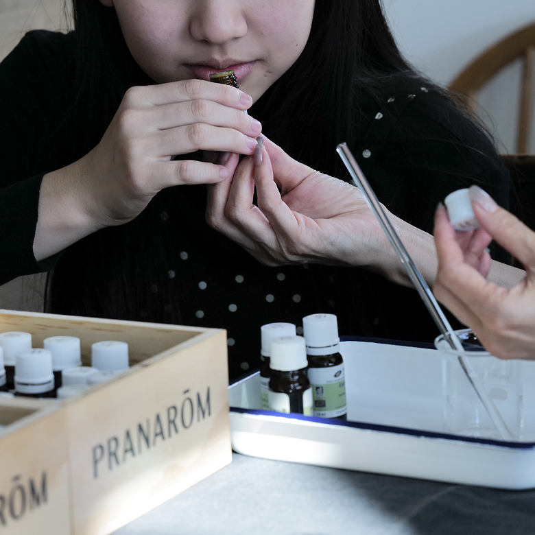 女の子に精油を差し出している。女の子はその精油の香りを嗅いでいる。テーブルの上にはたくさんの精油やガラスのビーカーが置いてある